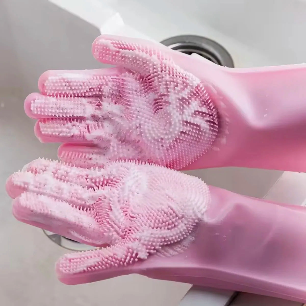A pair of pink silicone dishwashing gloves with textured scrubbers are shown in a close-up against a white sink.