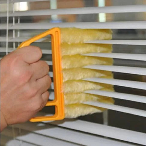 A hand is holding a yellow multi-functional cleaning brush with yellow bristles, positioned near white horizontal slats of bl