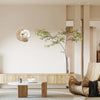 Une horloge murale au design lunaire moderne suspendue sur un mur clair au-dessus d'un meuble en bois et d'un arbre en pot.