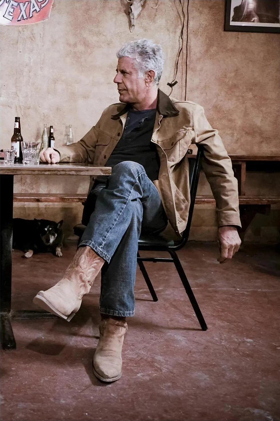Ein mittelalter Mann in einem beigen Jackett und Jeans sitzt auf einem Stuhl in einem rustikalen Interieur.