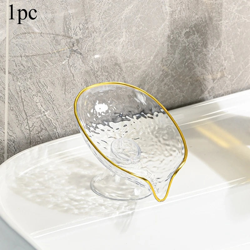 Clear oval plastic soap dish with gold rim and drainage holes resting on a white surface.