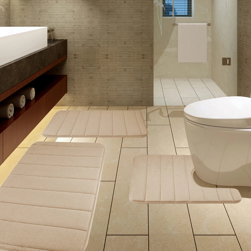 Ein Set aus drei beigen Memory-Schaum-Badematten auf einem gefliesten Boden neben Toilette und Waschtisch.