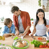 A woman and a boy are preparing food on a wooden table using a clear microwave splatter cover with a water steamer.