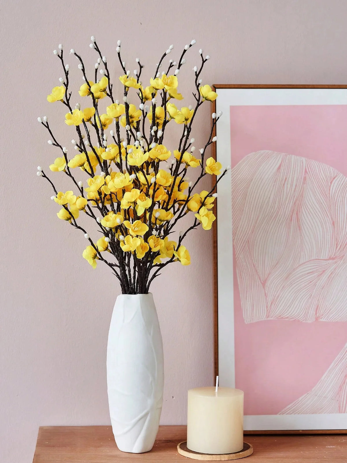 Eine Vase mit künstlichen gelben Pflaumenblütenzweigen neben einem gerahmten rosa Aquarellgemälde.