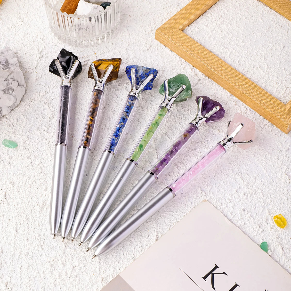 A collection of natural crystal pens featuring various colors including amethyst, quartz, and jade, resting on a white surfac
