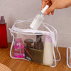 A hand is pouring liquid into a clear plastic travel makeup organizer bag containing three cosmetic bottles.