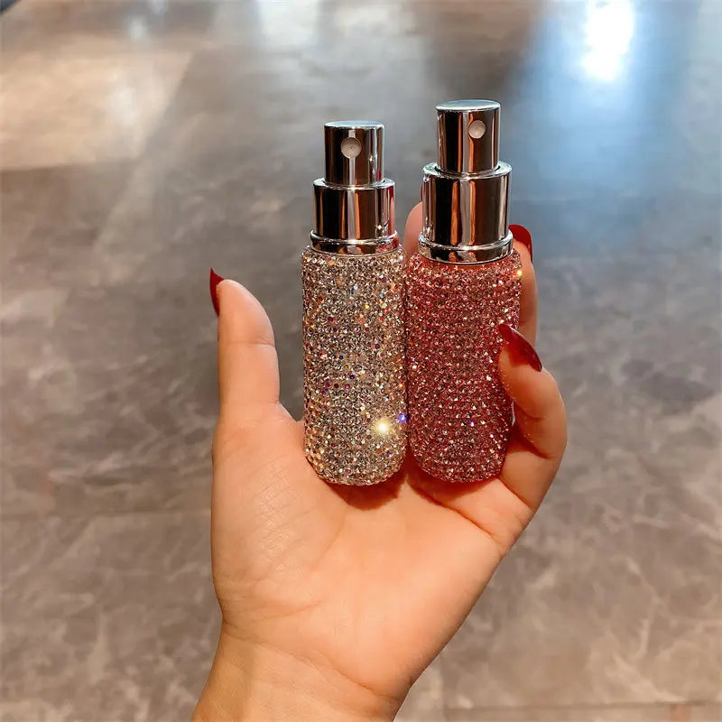 A hand holds two glass perfume spray bottles filled with sparkling pink and gold liquid.
