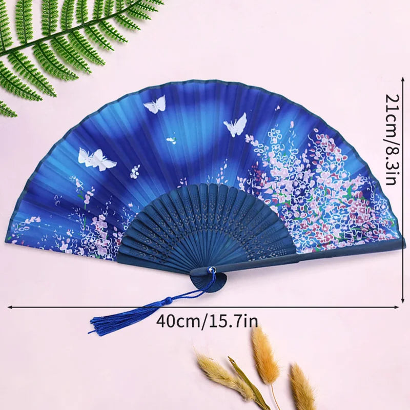 A blue and purple lace fabric folding hand fan displayed on a white background with a green fern leaf nearby.