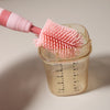 A pink silicone brush is shown dipping into a clear baby bottle containing a light yellow liquid.