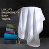 A white luxury embroidery bath towel draped over a stand, with stacked colored towels in the background.