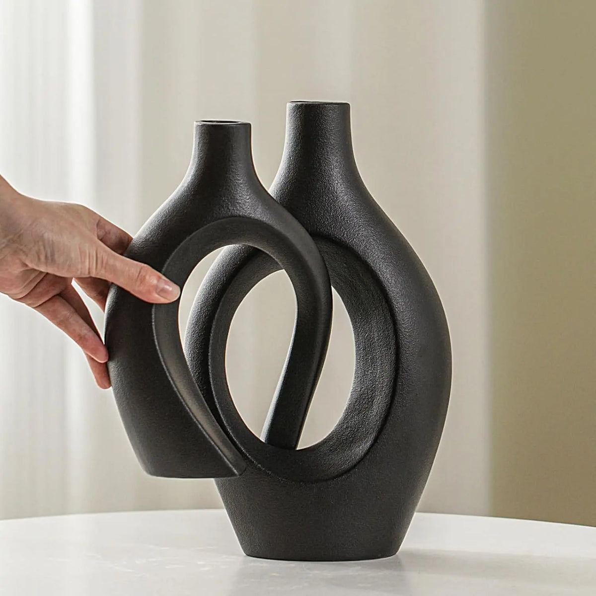 A hand holds a matte black ceramic vase with an organic, looped shape against a light background.