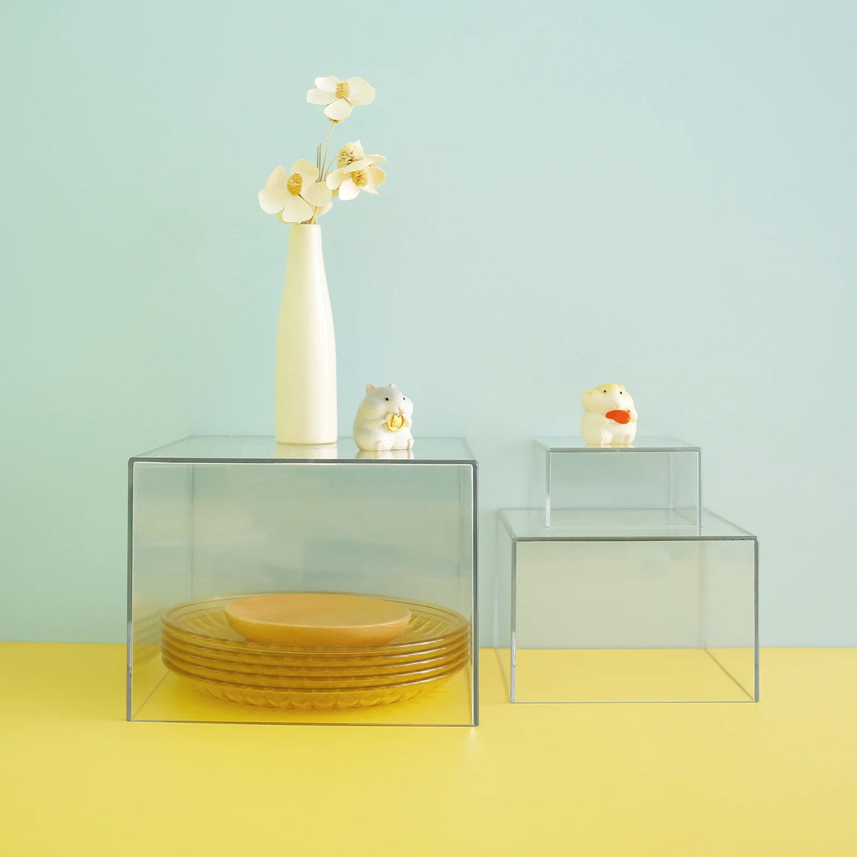 Clear acrylic stackable buffet riser displaying a white vase with yellow flowers and a small ceramic animal figurine.