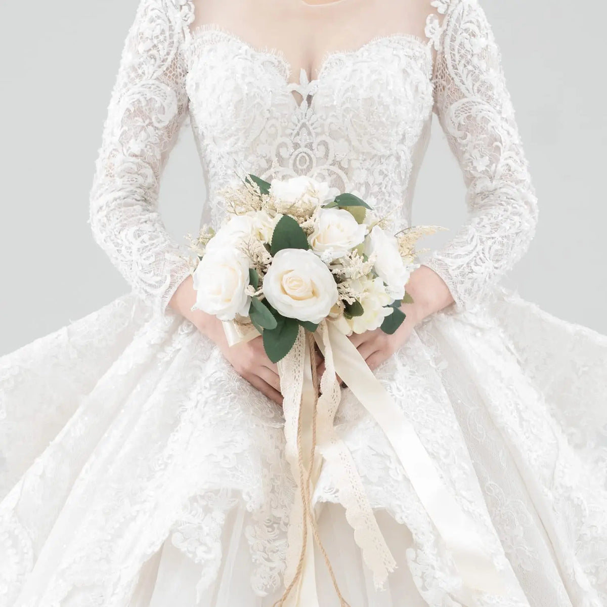 A bride wearing a white lace wedding dress holds an elegant bouquet of white and cream roses.