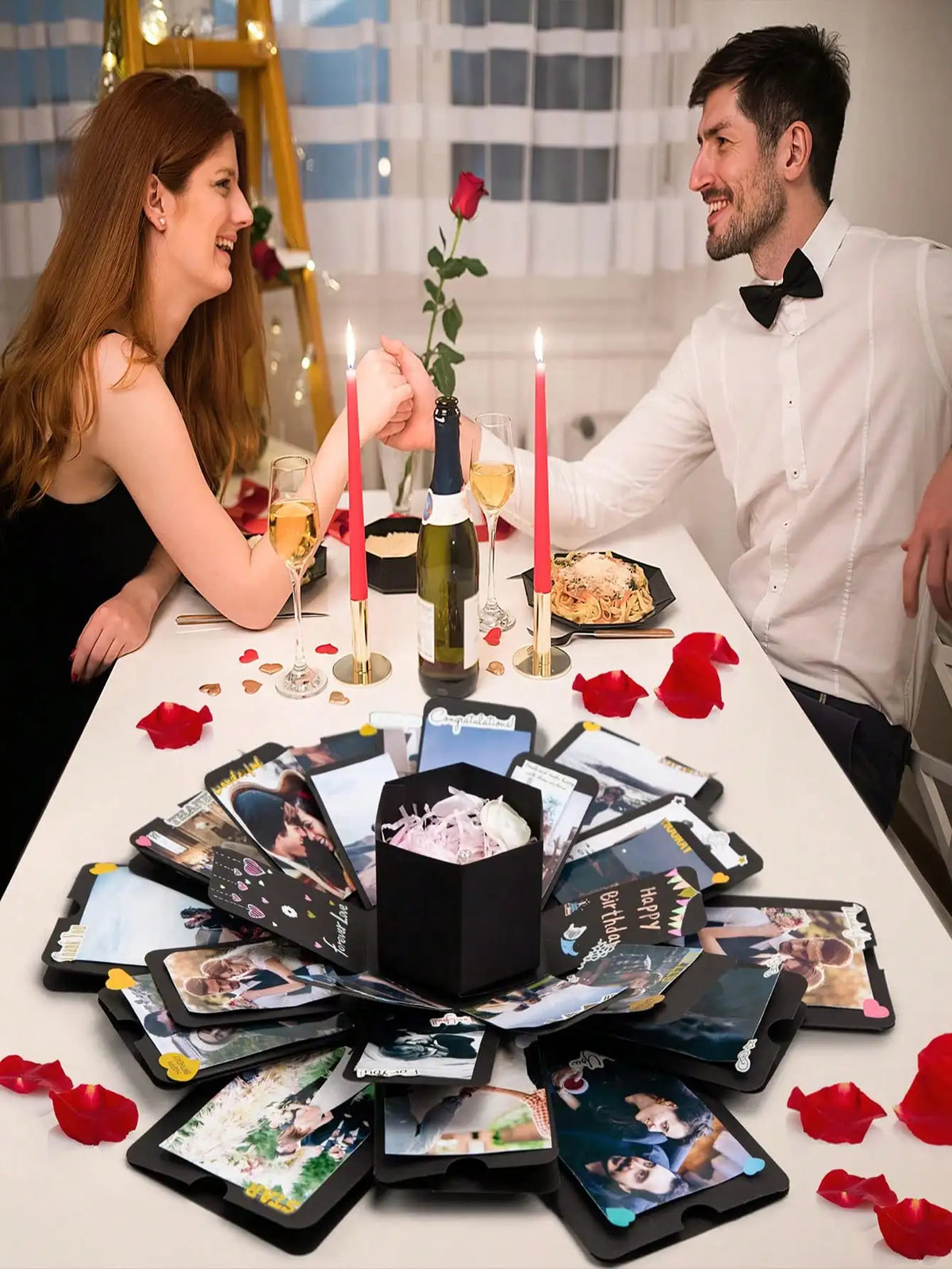 Ein Paar betrachtet eine DIY-Fotoalbumbox mit Drucken und roten Rosenblättern auf einem weißen Tisch.