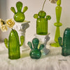 Collection de vases et sculptures en cactus en verre vert vif, présentés sur un socle blanc et de la fourrure blanche.