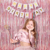 A smiling young girl wearing a pink ruffled top and skirt stands in front of a pink and gold tinsel fringe curtain backdrop w