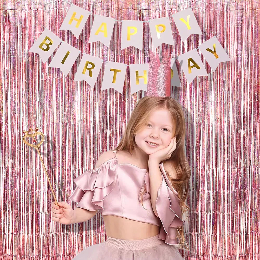 A smiling young girl wearing a pink ruffled top and skirt stands in front of a pink and gold tinsel fringe curtain backdrop w