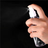 A hand is spraying a clear liquid from a white refillable portable spray bottle against a dark background.