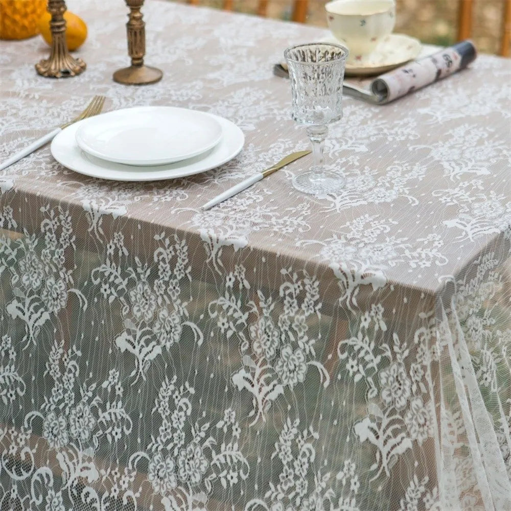 A table set with white plates, silverware, and a glass, covered by a sheer lace retro tablecloth.