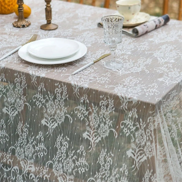 A table set with white plates, silverware, and a glass, covered by a sheer lace retro tablecloth.