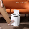 A white touchless sensor trash can with a lid is placed on a brown carpet in front of an orange sofa.