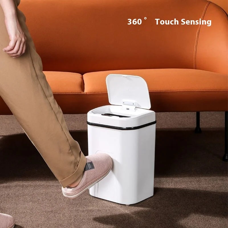 A white touchless sensor trash can with a lid is placed on a brown carpet in front of an orange sofa.