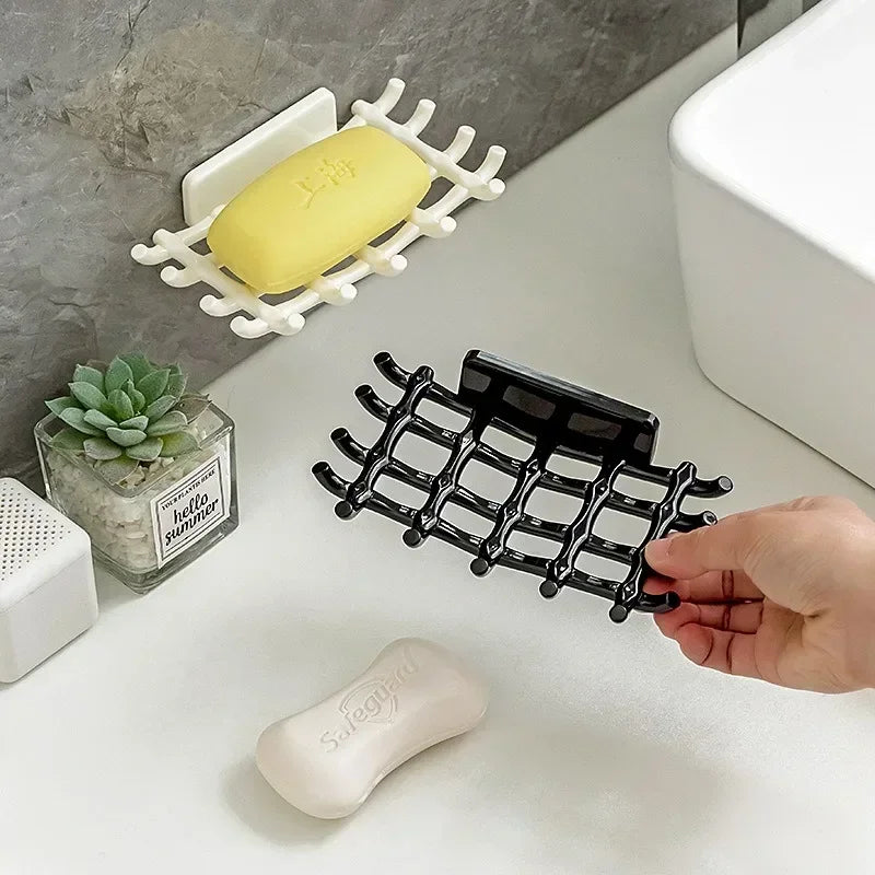 A hand holds a black wall mounted soap dish rack with multiple holes, next to a yellow soap bar and a white toilet.