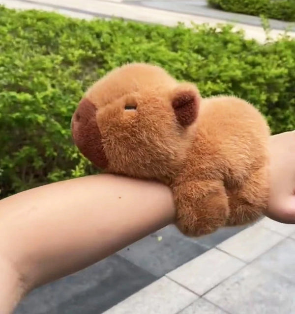 A small, fluffy capybara-shaped wristband bracelet is shown on a person's arm against a green background.