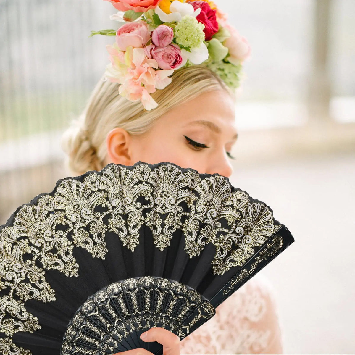 A woman holds a black and gold patterned folding fan while wearing a floral crown.