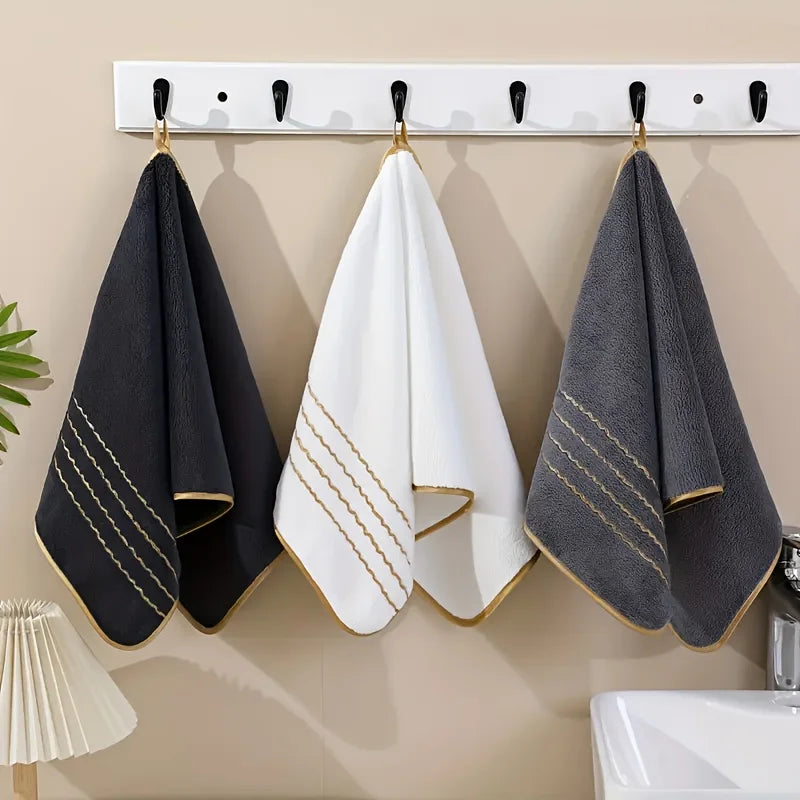 A set of three coral fleece towels in black, white, and gray with gold stripe details hanging on a white rack.