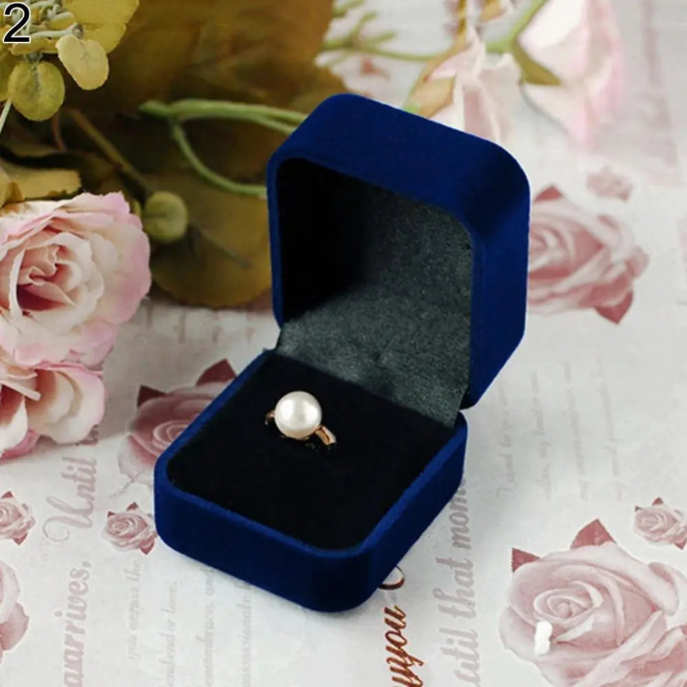 A dark blue velvet jewelry box open to display a single gold ring on a white background with pink roses.