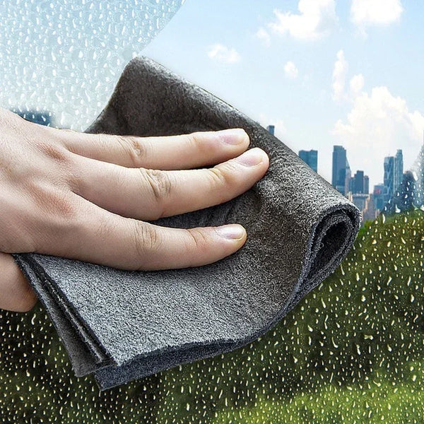 A hand is holding a dark gray microfiber cleaning cloth against a blurred background of greenery and city skyline with rain.