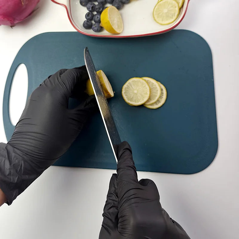 A hand wearing a black nitrile glove is using a knife to cut a lemon slice on a dark cutting board.