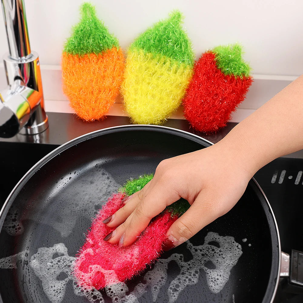 A hand is using a red strawberry scrubber sponge to clean food in a black frying pan.