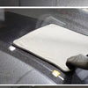 A microfiber car cleaning and drying towel is placed on a dark car surface, with a small yellow cleaning pad visible nearby.