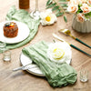 A rustic table setting featuring a green sheer fabric runner, white plates, and floral arrangements on a wooden surface.