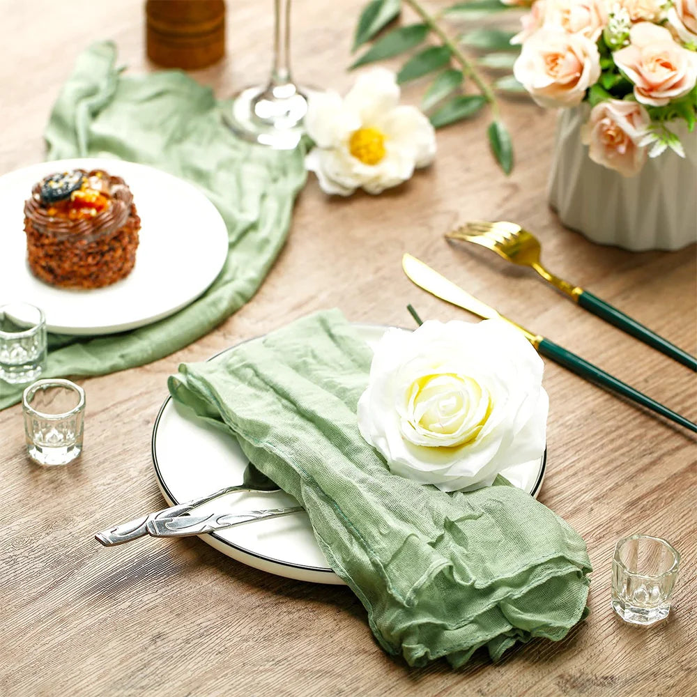 A rustic table setting featuring a green sheer fabric runner, white plates, and floral arrangements on a wooden surface.