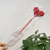 Une fleur en forme de cœur rouge en coton est exposée dans une gaine en plastique transparent sur un fond de plante verte.