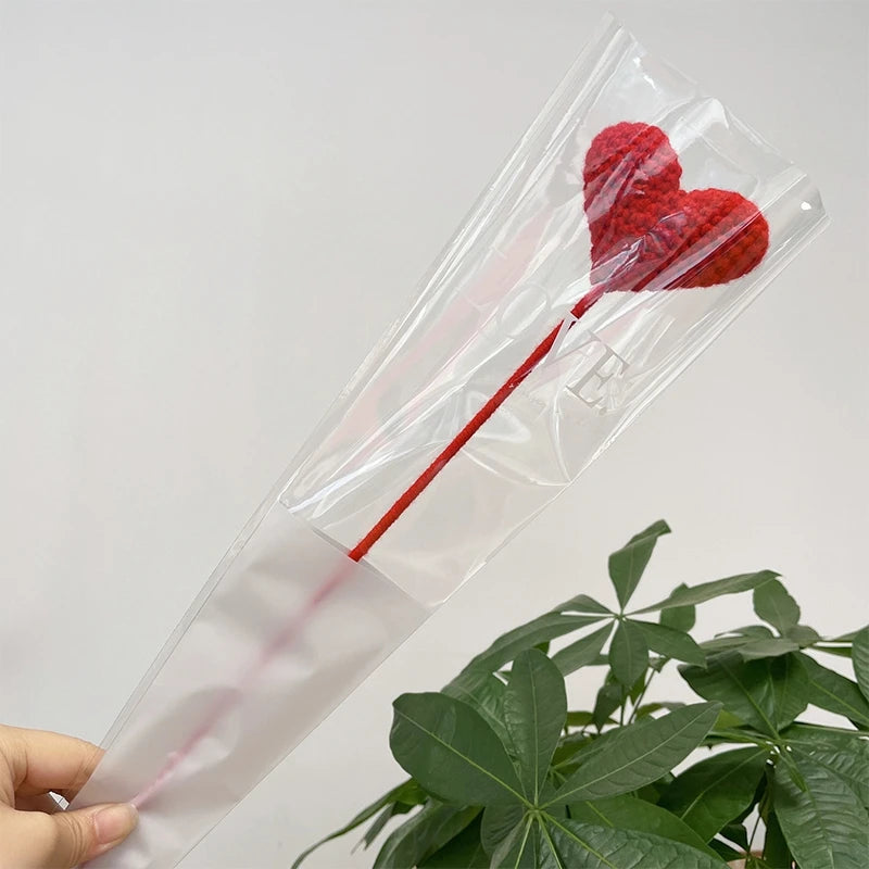 Une fleur en forme de cœur rouge en coton est exposée dans une gaine en plastique transparent sur un fond de plante verte.