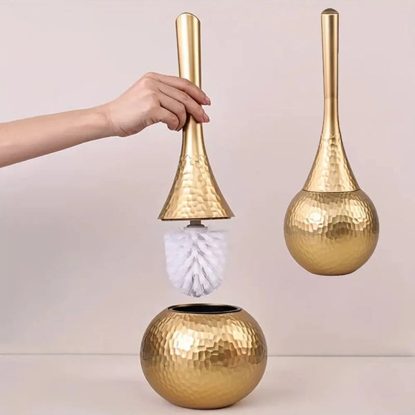 A hand holds a gold-colored long-handled toilet cleaning brush with a spherical head and a matching golden base.