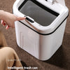 A white intelligent trash can with a lid is shown being opened by a hand on a brown carpet.