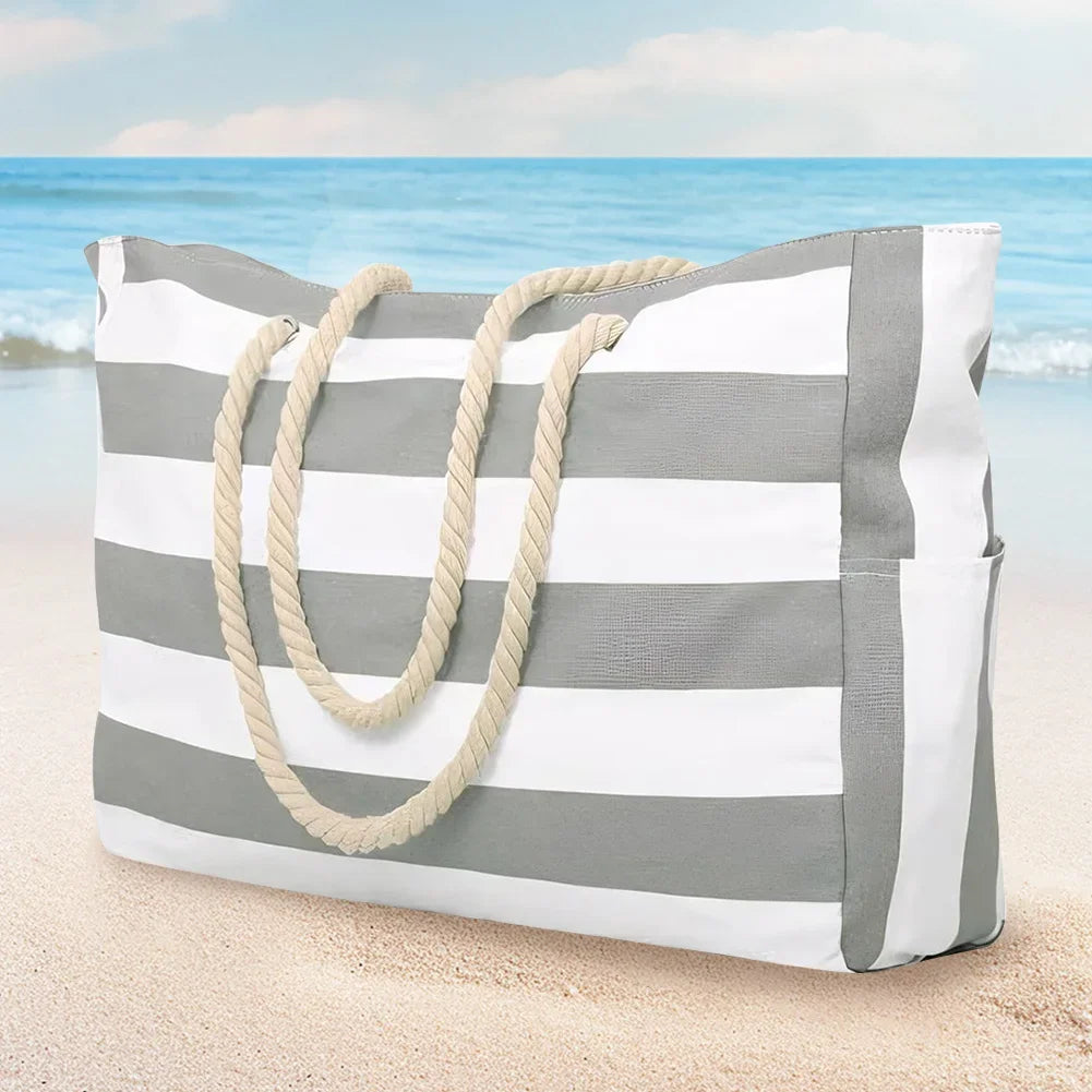 Weiße und grau gestreifte wasserdichte Strandtasche mit Naturfaser-Seilgriff am Sand nahe dem Ozean.