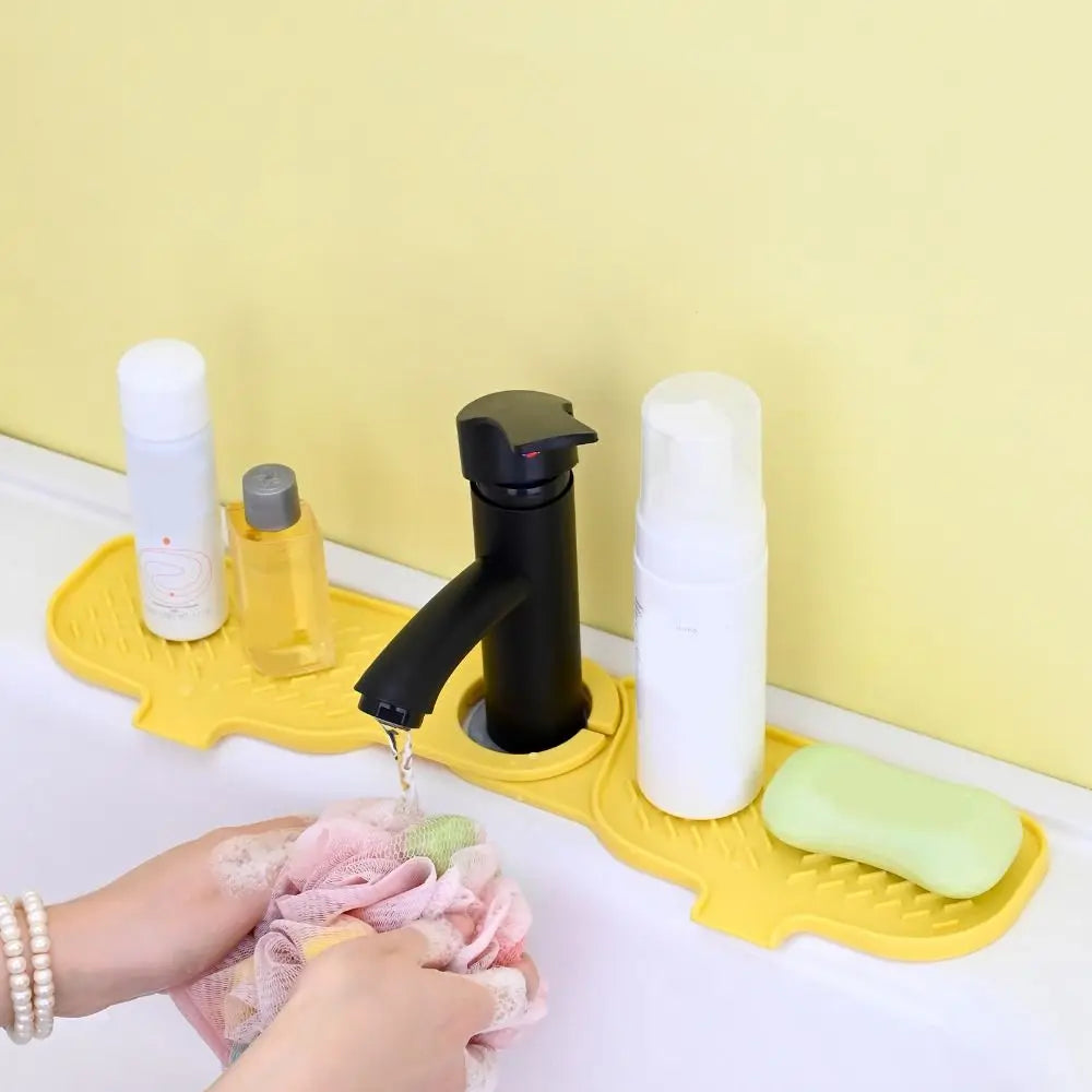 A yellow silicone mat holds various white and yellow liquid bottles and a black dispenser, with a hand holding a pink cloth.