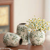 A white ceramic bud vase with green floral pattern sits on a wooden surface next to a pearl necklace.