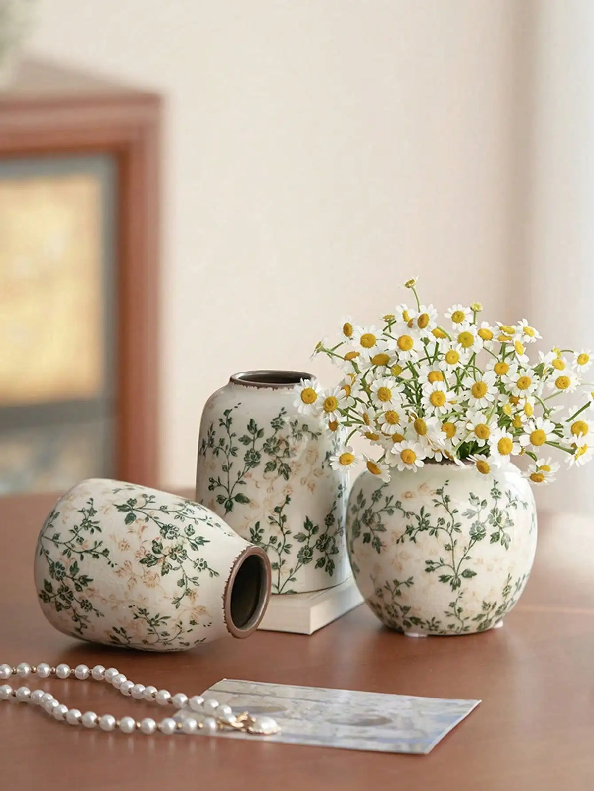 A white ceramic bud vase with green floral pattern sits on a wooden surface next to a pearl necklace.