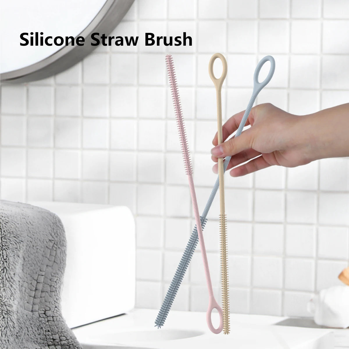 A hand is holding two silicone straw cleaning brushes, one pink and one light blue, against a white tiled background.